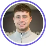 A headshot of a smiling short haired brunette man wearing a grey jumper with a purple ring around the image.