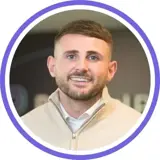 A headshot of a smiling brunette man with stubble wearing a beige jumper and white shirt underneath with a purple ring around the image.