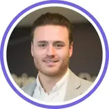 A headshot of a smiling brunette man with a stubble beard wearing a white shirt and light grey blazer with a purple ring around the image.