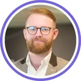 A headshot of a smiling ginger man with a full beard and glasses wearing a shirt and blazer with a purple ring around the image.