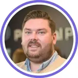 A headshot of a smiling brunette man with stubble wearing a light brown jumper and a blue and white chequered shirt under with a purple ring around the image.